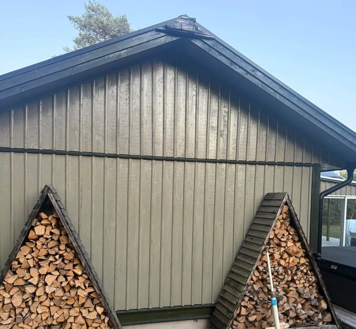 Gavel med olivgrön stående panel och svart vindskiva, med två vedstaplar framför